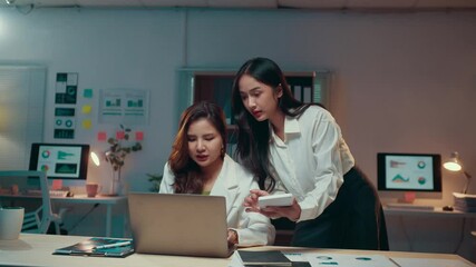 Poster - Two young businesswomen are working late at the office. One is sitting at a desk using a laptop while the other stands beside her, using a calculator and reviewing paperwork