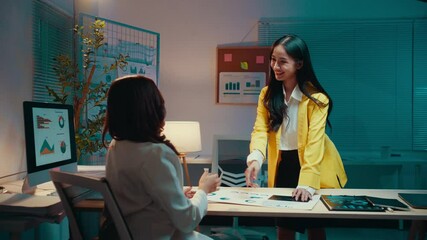 Poster - Two young businesswomen shaking hands after finalizing a contract in a modern office setting at night, celebrating a successful partnership and demonstrating teamwork and collaboration