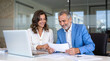 © Sachin - Cheerful middle aged business professionals, Hispanic woman in white blouse and senior man in blue blazer, reviewing documents together in modern office
