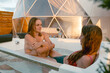 © Alina Hvostikova/Stocksy - Two women relax in a hot tub near a geodesic dome at sunset