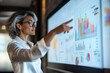 © Olena - Portrait of female professional worker in a white shirt and glasses is touching a large screen with data curves, graphs, and tables. Woman standing near a large digital display in office.