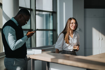 Wall Mural - Two business workers enjoying a break in a modern office setting. They are smiling and engaged, showcasing a relaxed, multicultural work environment. Ideal for concepts of teamwork and workplace