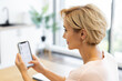 © sofiko14 - Caucasian woman in her 30s shopping for cosmetics on online delivery website using smartphone. She sits in bright, modern home environment, focusing on her phone screen.