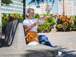 © luciano - Smiling relaxed tourist woman enjoys a sunny day sightseeing Santa Cruz of Tenerife sitting in a bench looking at map indications, city life in background