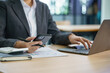 © Songsak C - Close-up image of a businesswoman holding a smartphone and typing on a laptop computer on the table.