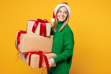 Wall Mural - Merry young woman wear green casual sweater Santa hat posing hold stack of present boxes with gift ribbon bow isolated on plain yellow background. Happy New Year celebration Christmas holiday concept