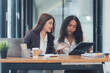 © Kritdanai - Collaborative Businesswomen Reviewing Project: Two diverse businesswomen, one Asian and one African-American, collaborate intensely over a tablet in a modern office setting.
