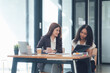 © Kritdanai - Collaborative Teamwork: Two smiling businesswomen engage in a productive discussion, reviewing documents and collaborating on a project in a modern, well-lit office setting.