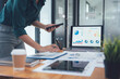 © Kritdanai - Analyzing Data: A business professional examines financial data on a laptop and tablet, surrounded by other work essentials in a modern office setting.