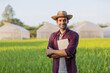 © kamonrat - Confident Farmer Standing in a Lush Green Field with Tablet, Smiling at Camera, Showcasing Sustainable Farming and Modern Agricultural Practices with Greenhouses in Background