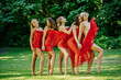 © Connect Images - Five women draped in red fabric strike playful poses on a sunny day with lush greenery in the background, USA