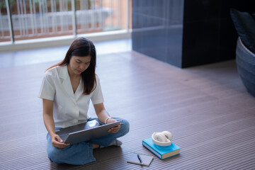 Wall Mural - Young asian woman sitting cross-legged on the floor, working remotely on a laptop while wearing headphones. A notebook rests nearby, creating a comfortable and focused workspace