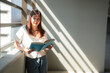 © Tj - Asian woman enjoying reading a book while standing by a large window, soaking in the warm, natural light that fills the room and creates a peaceful, serene atmosphere