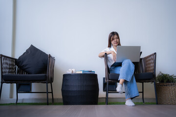 Wall Mural - Young asian businesswoman is working from home, sitting on a comfortable chair on her balcony, using a laptop and enjoying the natural light and fresh air