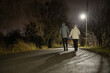 © StockMediaSeller - Couple holding hands while walking on a quiet road at night, illuminated by a streetlight, surrounded by bare trees and a serene atmosphere.