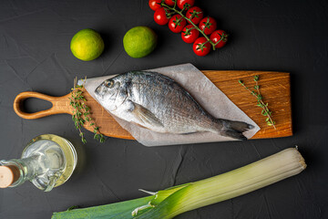 Wall Mural - Fresh, raw dorado fish on a wooden board on a black background with leeks, lime, olive oil and cherry tomatoes on the table ready for cooking