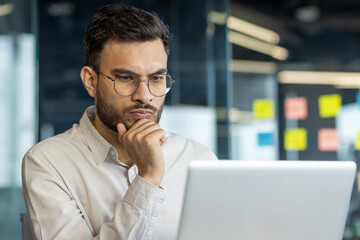 Wall Mural - This image features a man, immersed in work, thoughtfully analyzing data on his laptop in an office setting. The professional setting emphasizes focus, concentration, and decision-making aspects.