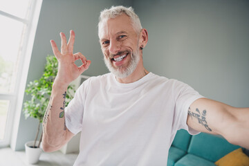 Wall Mural - Energetic mature man in a white t-shirt smiling indoors and making an okay gesture with his tattooed arm, enjoying a healthy