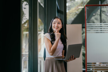 Fototapete - Young asian businesswoman holding laptop and thinking new ideas while looking away, touching her face with hand and smiling in modern office