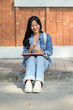 © bongkarn - A female college student sits on the edge of a footpath at her university, using her smartphone.