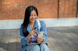 © bongkarn - A female college student sits on the edge of a footpath at her university, using her smartphone.