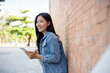 © bongkarn - Smiling female student stands leaning against a brick building wall outside, holding her smartphone.
