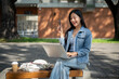 © bongkarn - A positive Asian female college student sits on a bench at her university, using her laptop to work.