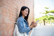 © bongkarn - A smiling female student stands leaning against a brick building wall outside, chatting on her phone