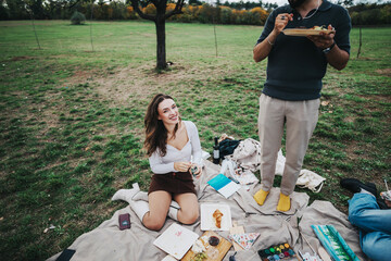 Canvas Print - A cheerful woman sitting on a picnic blanket outdoors, surrounded by food and art supplies, enjoying a relaxing day under a tree with a companion.