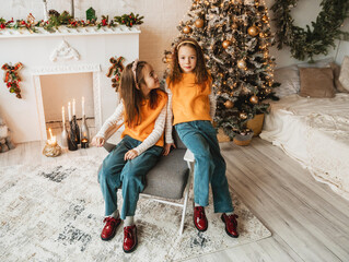 Wall Mural - Two adorable sisters in matching outfits pose for a Christmas portrait. They sit near a decorated fireplace and Christmas tree, creating a heartwarming holiday scene