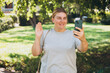 © mdyn - Hi. Female taking a selfie photo using her mobile phone in the public park. Woman in t-shirt recording video, talking on online call and relaxing outdoors. Urban people concept.
