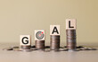 © Supatman - Financial Goals and Savings Concept. Wooden blocks spelling 'GOAL' rest on stacks of coins, with a target icon representing financial planning, savings, achieving monetary objectives, business growth,