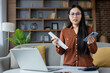© Liubomir - Asian woman holding receipt, phone while managing expenses in home setting. Expressing concern over financial issues with laptop on table. Business, finance, communication.