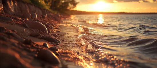 Wall Mural - Serene shoreline at sunset with gentle waves lapping against the sandy beach and glistening pebbles reflecting warm sunlight.