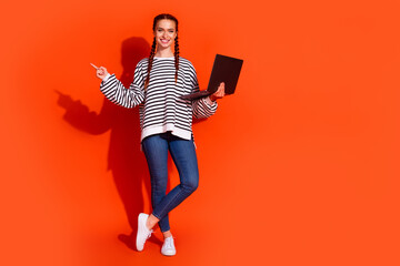 Young woman with red hair in striped pullover holding laptop on orange background