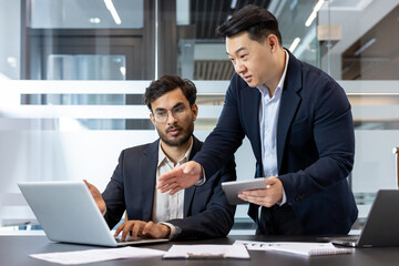 Wall Mural - Two business professionals engage in a collaborative discussion in a modern office environment. One stands holding a tablet, while the other works on a laptop. The setting highlights teamwork.