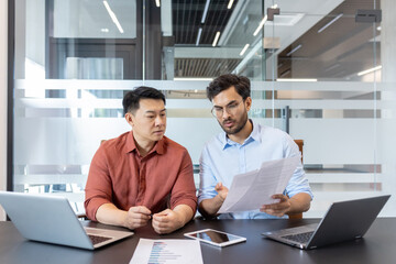 Wall Mural - Two professionals engage in a discussion over important documents in office. Laptops and a phone are on the table, suggesting a tech-driven work environment. Collaboration and focus are evident.