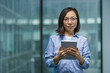 © Liubomir - Asian woman confidently holds a tablet in a modern office. She appears professional, wearing glasses and a blue shirt, symbolizing success and technology in a business setting.