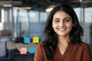 Wall Mural - A confident young businesswoman in a modern office, smiling and posing for the camera. Her expression exudes professionalism and positivity, ideal for business presentations.