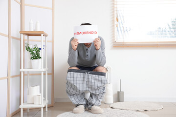 Wall Mural - Man holding paper with word HEMORRHOID while sitting on toilet bowl in restroom