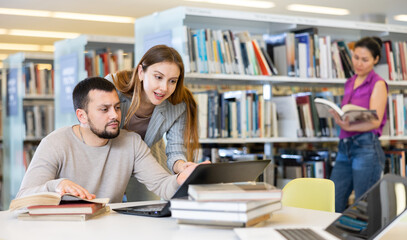 Young guy and girl students studying in the university library are preparing an abstract on a laptop and looking for sources ..of information in books