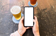© Ibai Acevedo/Stocksy - Hands using white screen phone with beers at table