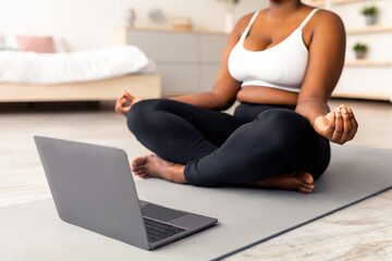 Wall Mural - Plus size black woman having online meditation or yoga class, sitting in lotus pose next to laptop at home, cropped view. Unrecognizable overweight young lady doing breathing exercises indoors