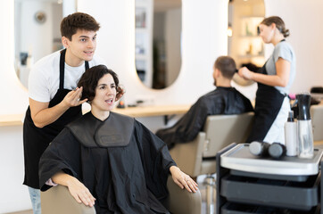 Wall Mural - Young guy hairdresser stylist gives haircut to adult woman client in beauty salon