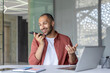 © Liubomir - A cheerful business professional uses his smartphone for voice messaging in a modern office setting. The open laptop and notebook on the desk suggest a productive and efficient workspace.