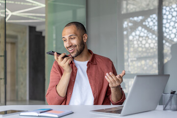 A cheerful business professional uses his smartphone for voice messaging in a modern office setting. The open laptop and notebook on the desk suggest a productive and efficient workspace.