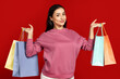© Prostock-studio - A young Caucasian woman is smiling brightly while holding shopping bags in each hand. She appears joyful and content, showcasing vibrant colors of the bags against a striking red background.
