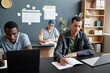 © Seventyfour - Medium shot of adult multiracial man concentrated on writing task sitting at desk in study group with other students attending language courses class