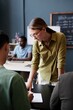 © Seventyfour - Vertical shot of young female teacher assisting student in completing writing task while standing at desk during lesson in English language school, copy space