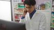 © Krakenimages.com - A young, handsome hispanic man in a white lab coat talks on the phone inside a well-stocked pharmacy, standing attentively among shelves filled with various medicinal products.